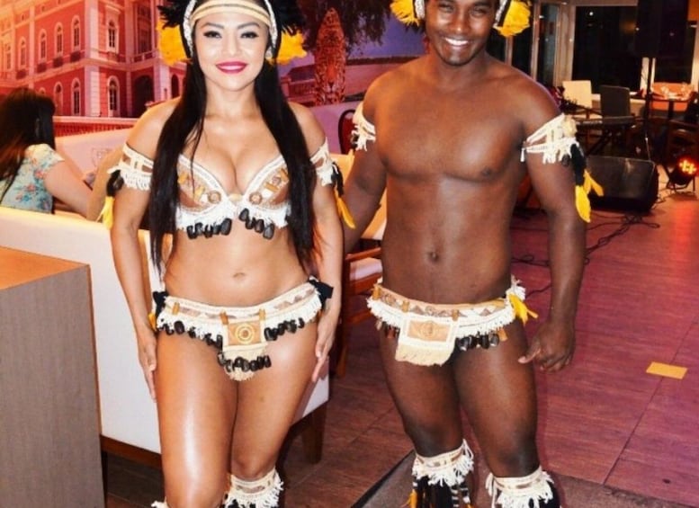 A man and a woman in traditional attire with beads and feathers, standing indoors with a red carpet and a building in the background. 1