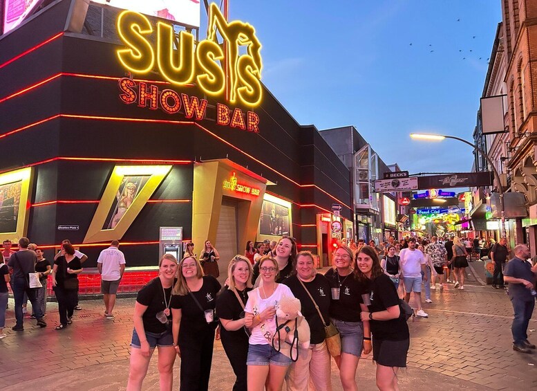 A group of people posing in front of a neon-lit bar named 'SUS's SHOW BAR' with a bustling street scene in the background. 2
