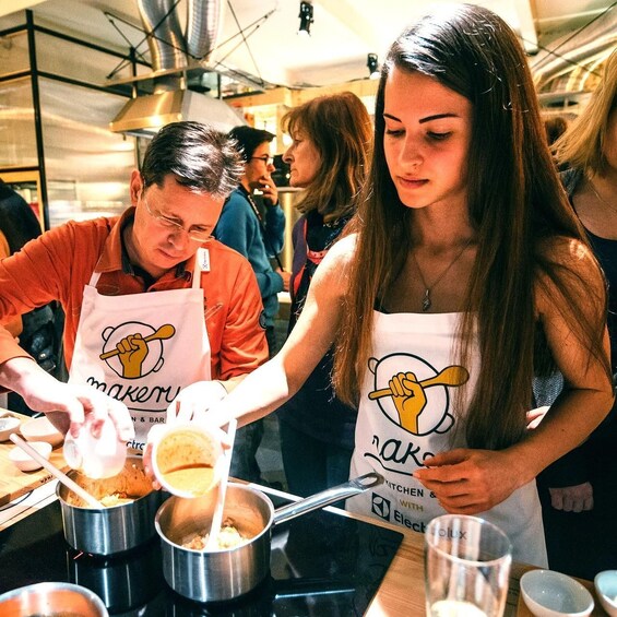 Two people wearing aprons with a kitchen logo, preparing food in a professional kitchen environment. 2