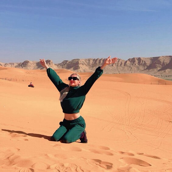 A person kneeling on a desert with arms raised, a vehicle in the background, and sand dunes. 1