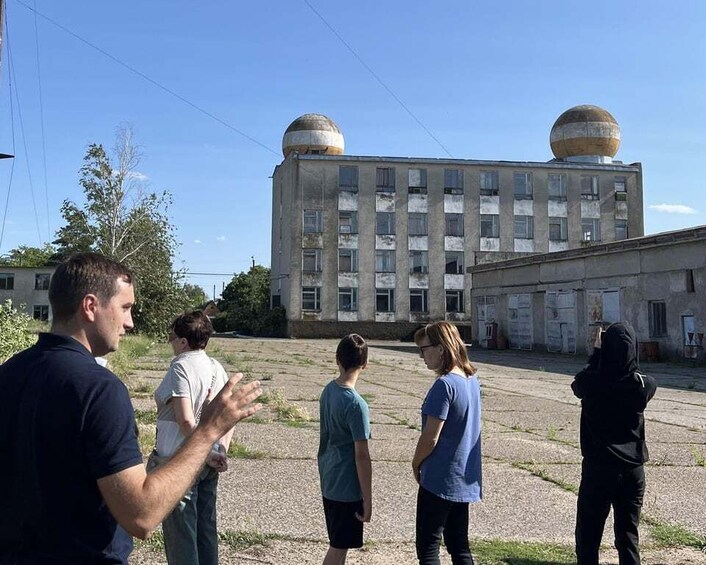 A group of people standing in an open area with a large, multi-story building featuring spherical structures in the background. 1