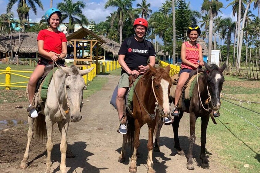 Three people riding horses on a dirt path with a thatched-roof structure and palm trees in the background. 2