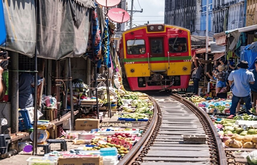 A train passing through a market with various goods displayed on the tracks. 3