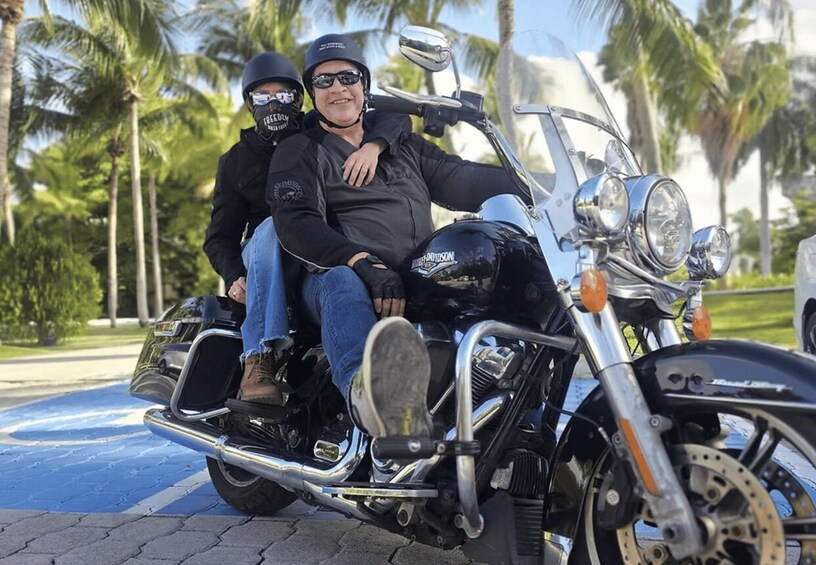 Två personer på en motorcykel med en Harley-Davidson-logotyp, parkerad på en asfalterad yta med palmer i bakgrunden. 4