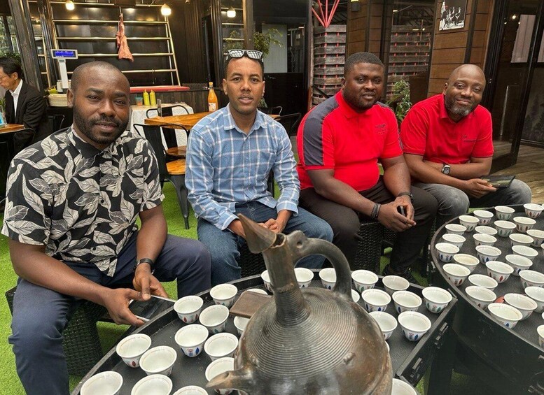 Four men seated around a table with multiple cups and a teapot. 1