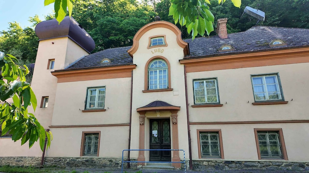 A two-story building with the name 'LUBO' on the facade, multiple windows, and a stone wall in front. 3