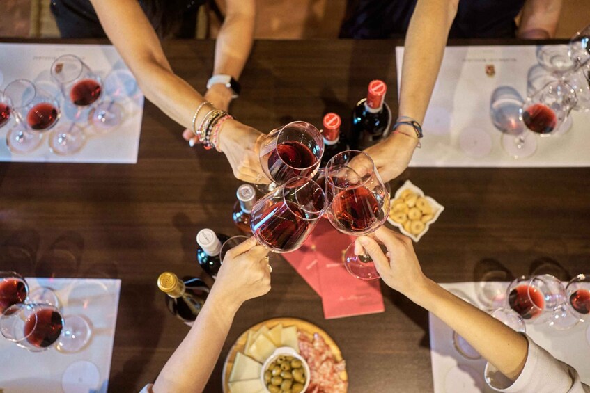A group of people toasting with wine glasses around a table with a cheese platter and bottles of wine. 1