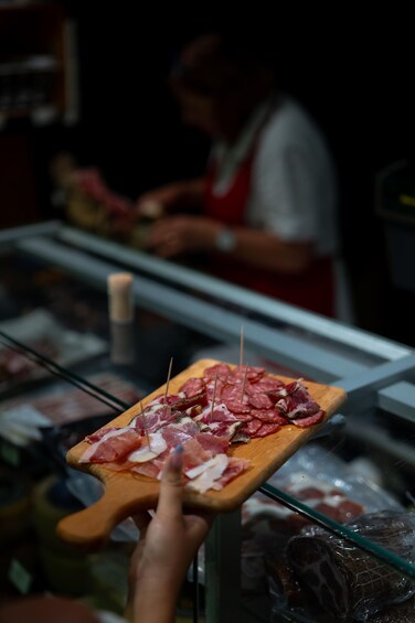A person holding a wooden board with sliced meat and toothpicks, in a meat shop. 1