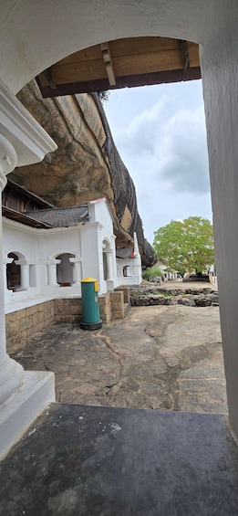 A stone pathway leading to a white building with an arched entrance, a green trash bin, and a large rock formation in the background. 5