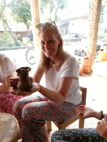 A person holding a clay pot, seated outdoors with a bicycle and pottery in the background. 5