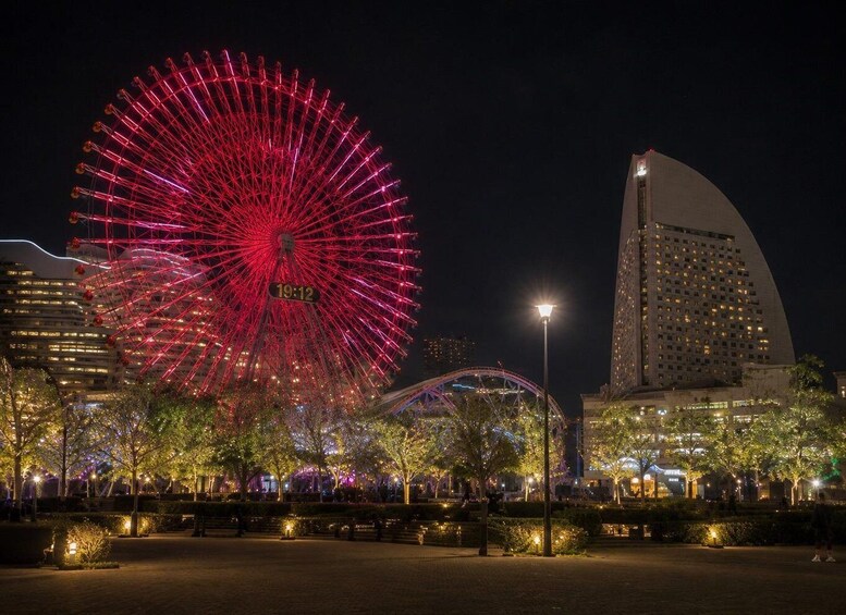 A large red Ferris wheel, a curved building, and a park with trees and benches at night. 2