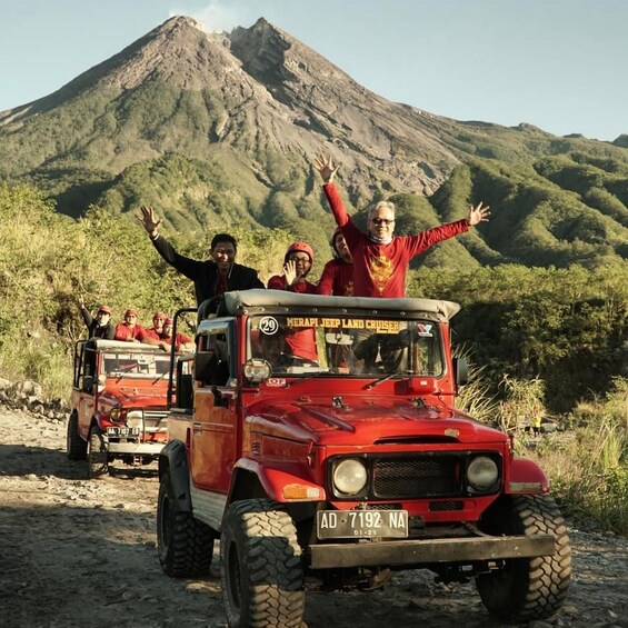 A group of people on a red jeep with 'MERAPI JEEP LAND CRUISER' written on it, driving on a dirt road with a mountain in the background. 4