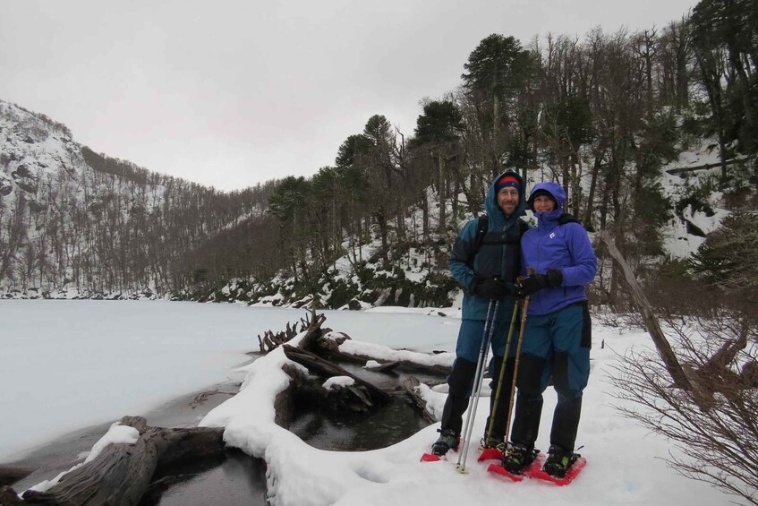 Two people standing on a snow-covered log over a frozen lake, surrounded by a forested mountainous landscape. 1