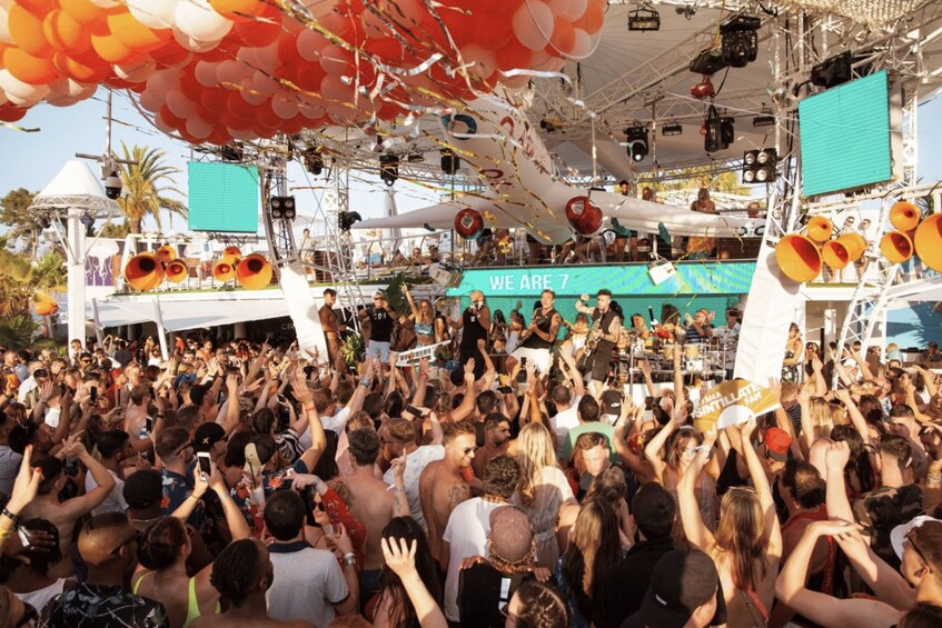 A crowded outdoor event with a stage and a banner that reads 'WE ARE 7'. 2