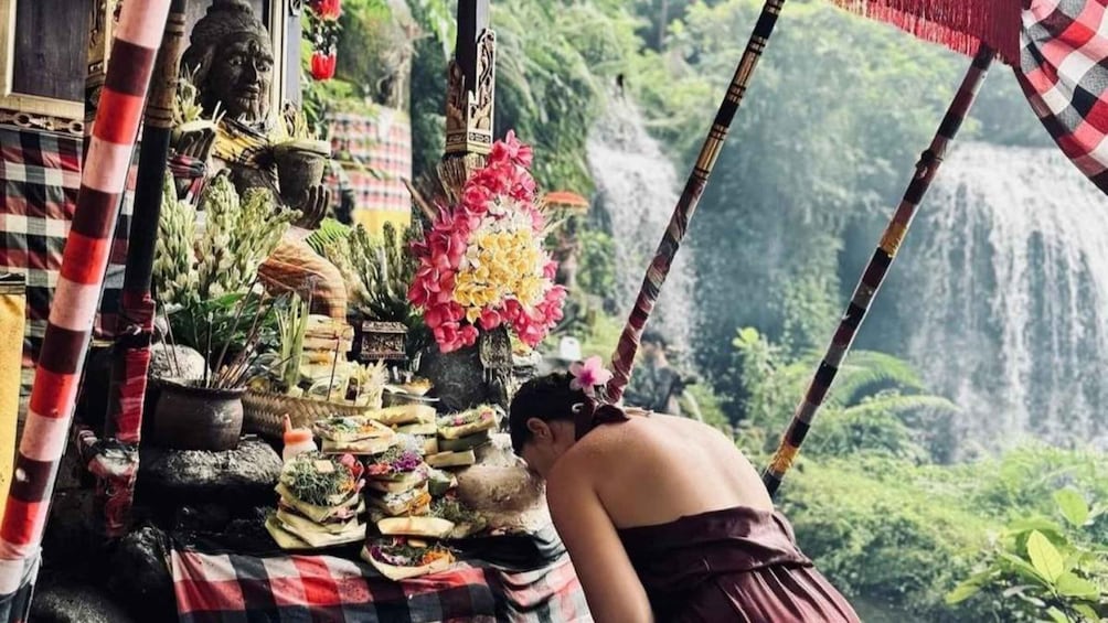 A person performing a ritual at a shrine with offerings, surrounded by lush greenery and a waterfall. 5