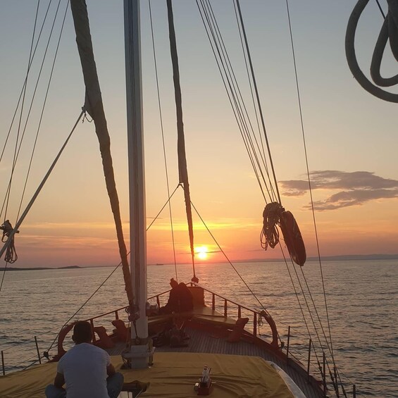 A sailboat at sunset with a person steering, a person relaxing on deck, and the ocean in the background. 5