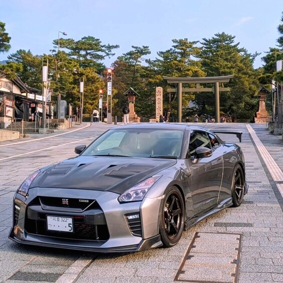 A silver sports car parked on a paved area with trees and a traditional Japanese torii gate in the background. 1