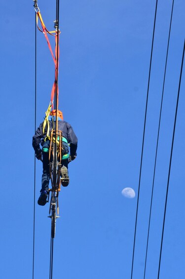 A person suspended in the air, attached to a cable system, with a clear blue sky and a visible moon in the background. 5