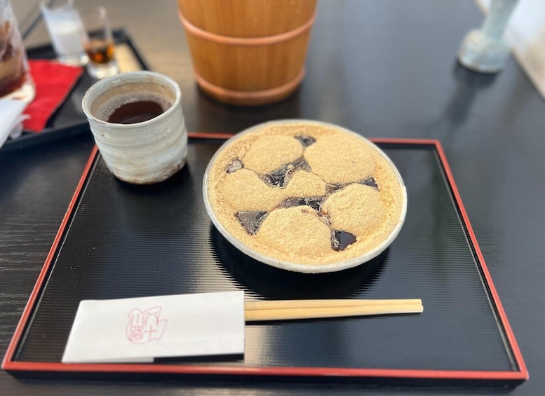A bowl of mochi with a red and black pattern, a small cup of green tea, and a pair of chopsticks on a black tray with a red border. 1