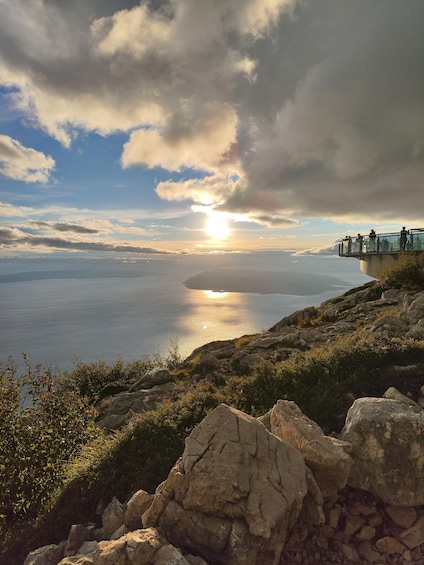 A coastal landscape with a rocky foreground, a glass railing overlooking the sea, and a sunset in the distance. 3