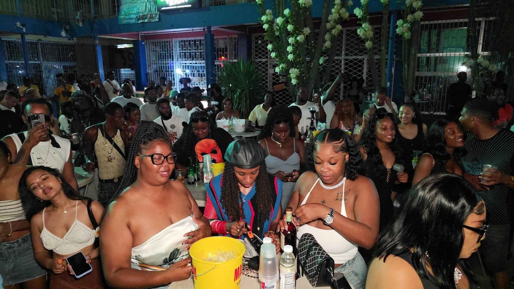 A crowded indoor event with people gathered around tables, some holding drinks and others engaged in conversation. 3