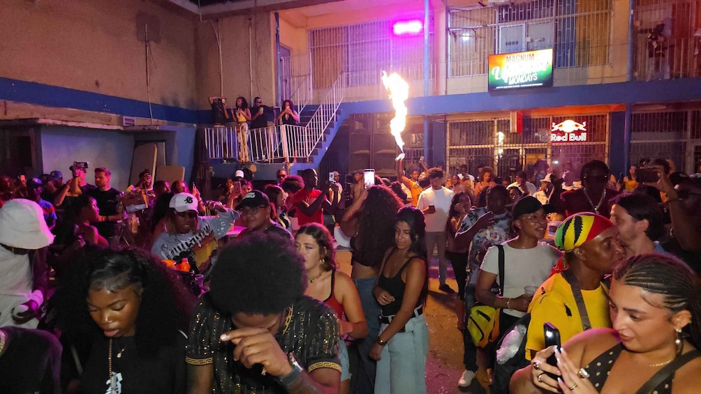 A crowded outdoor event with people gathered around a stage, a building with a sign reading 'MAGNUM,' and a 'Red Bull' logo visible. 4