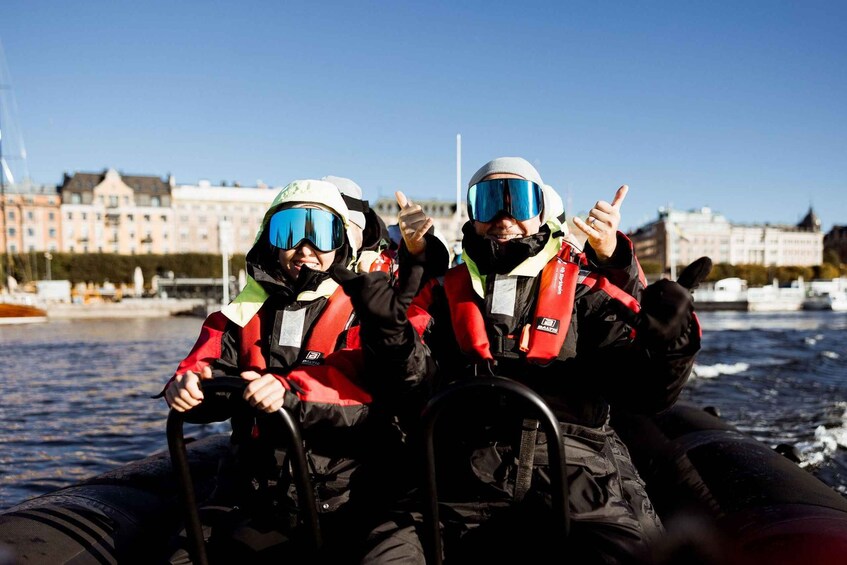 Two people in a boat wearing life jackets and helmets, with a cityscape in the background. 5