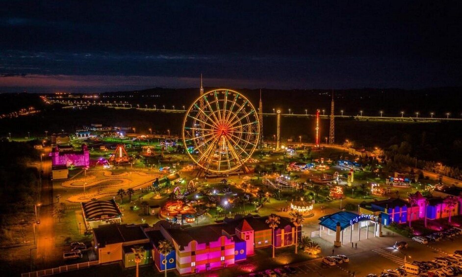 A brightly lit amusement park at night with a large Ferris wheel, colorful buildings, and a bridge in the background. 1