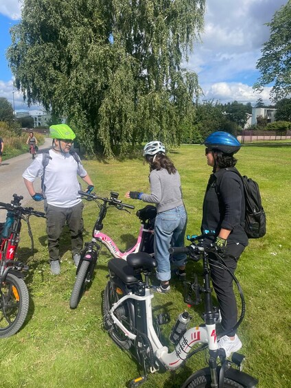 Three people with electric bikes on a grassy path, a tree, and a building in the background. 3