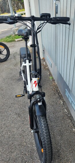 A stationary electric bicycle with a white frame and black components, parked on a paved surface next to a building. 2