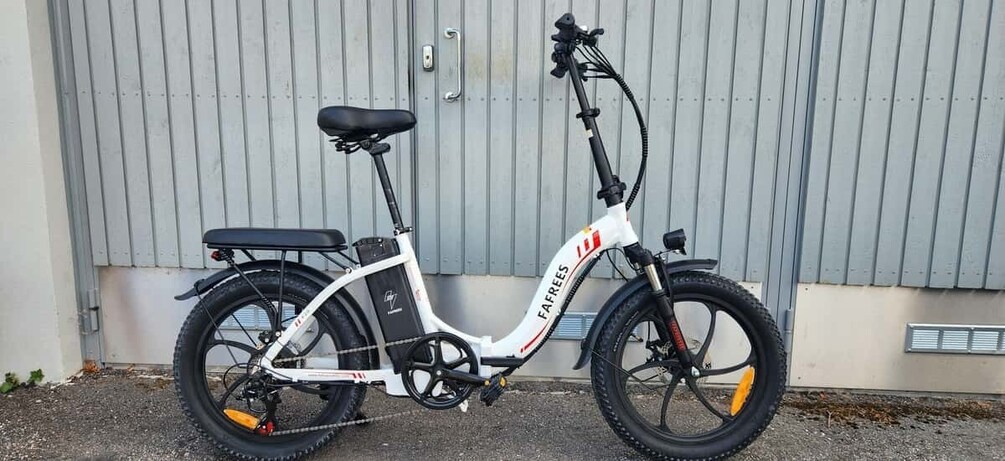 A white electric bicycle with black tires and a black seat, parked in front of a grey metal wall. 5