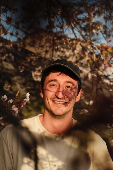 A person wearing a cap and a light-colored shirt, standing outdoors with trees and sunlight in the background. 1