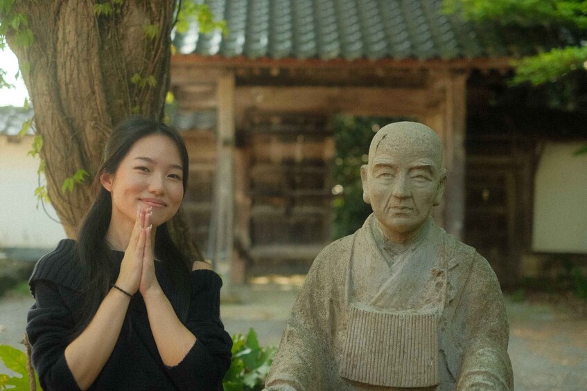 A person in black clothing standing next to a stone statue in a garden with a traditional building in the background. 3