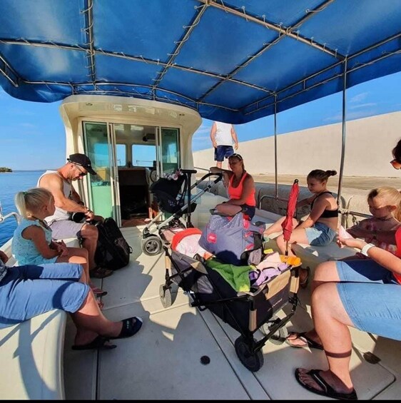 A group of people on a boat with a blue canopy, some sitting and others standing, near a sandy shore. 4