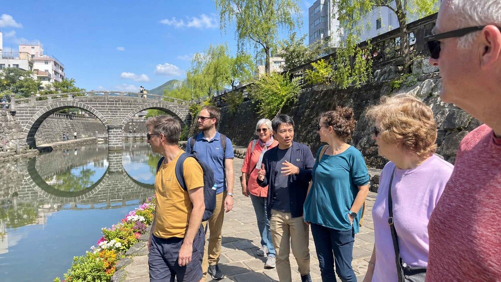 A group of people walking along a riverside path with a stone bridge and buildings in the background. 2