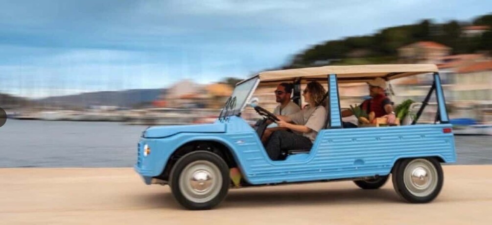 A blue vintage car with passengers, driving on a waterfront road. 1