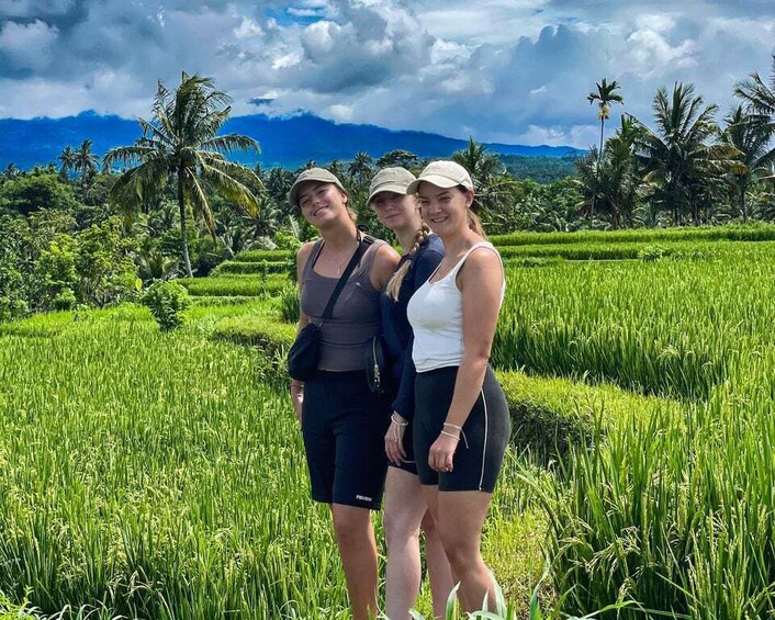 Three people standing in a lush green rice field with palm trees and mountains in the background. 2