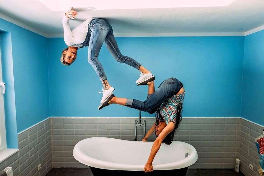 Two people performing an acrobatic stunt in a bathroom with blue walls and a white bathtub. 1
