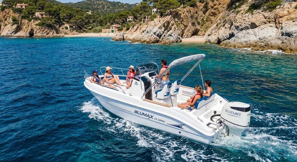A white motorboat named BLUMAX 23 OPEN on clear blue water with people on board, rocky coastline, and buildings in the background. 1