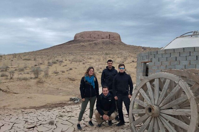A group of people in a desert landscape with a large mound in the background and a wooden wagon wheel in the foreground. 3