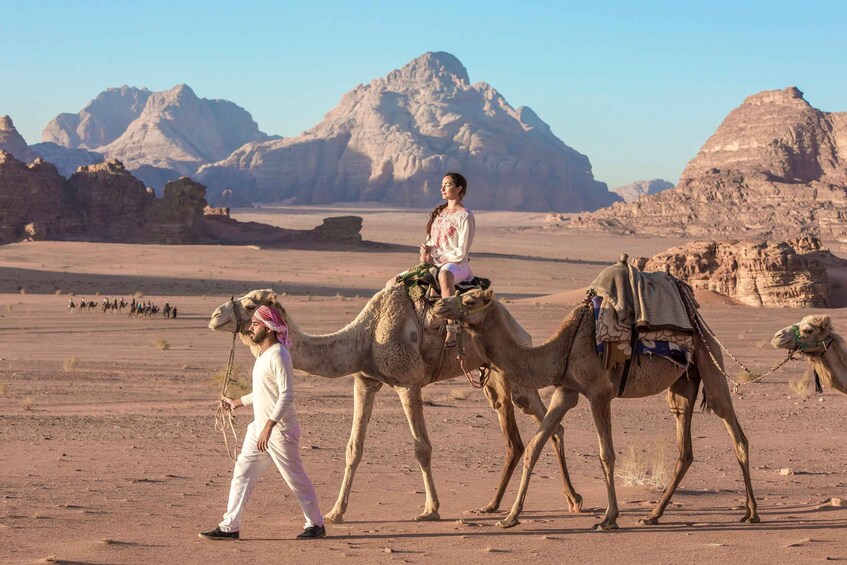 A desert landscape with rocky mountains in the background, a person leading camels, and a camel caravan. 2