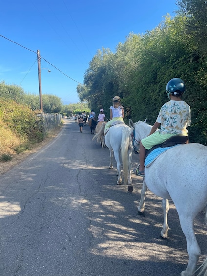 A group of people riding horses down a narrow road flanked by trees and vegetation. 1