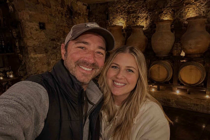 A person in a dark vest and cap, standing in front of stone walls with large pottery and wooden barrels. 1
