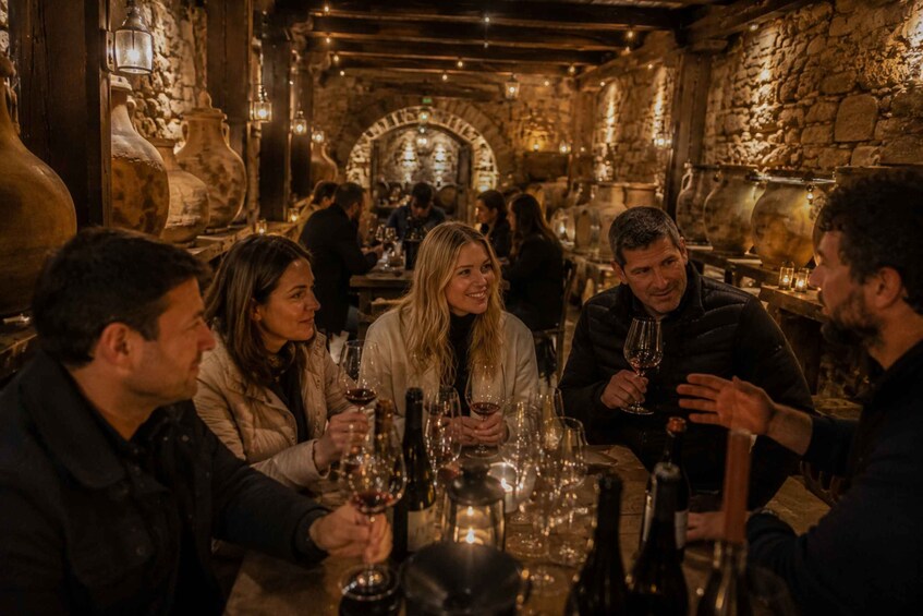 A group of people seated at a table in a dimly lit, stone-walled interior with large pottery vases. 2