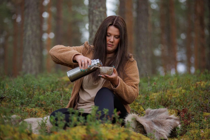 A person sitting on the ground in a forest, holding a metal container. 1