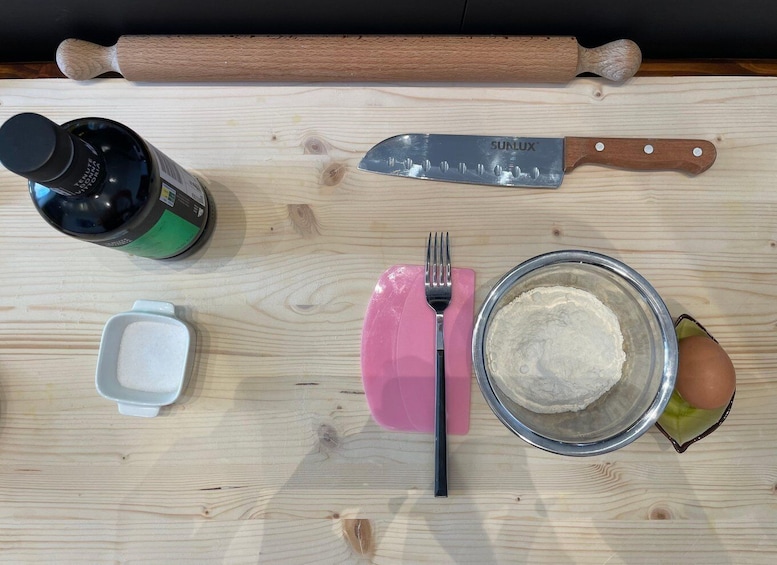 A wooden surface with a rolling pin, a knife, a bottle, a fork, a bowl of flour, a small dish of salt, and a peeled egg. 3