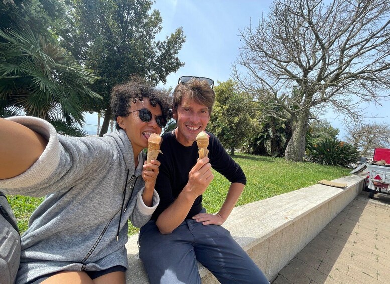 Two people sitting on a concrete ledge, holding ice cream cones, with trees and a clear sky in the background. 5