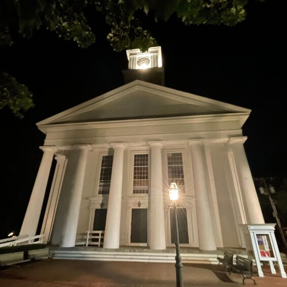A white neoclassical building with columns and a clock at the top, illuminated at night. 1