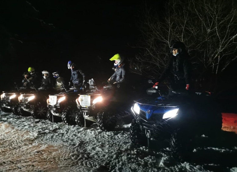A group of riders on ATVs with headlights on, navigating a snowy trail at night. 3