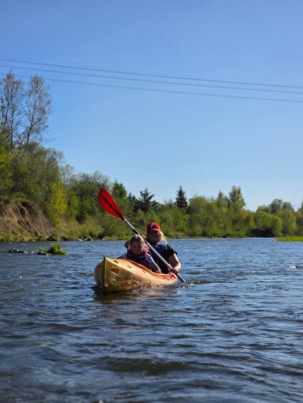 Two people kayaking on a river with trees and a clear sky in the background. 4